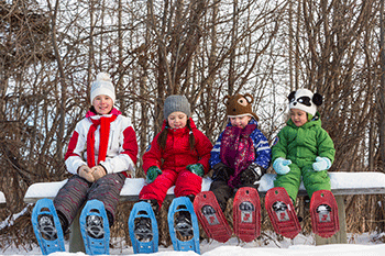 Enfants souriants assis sur un banc et des raquettes dans les pieds dans un boisé en hiver.