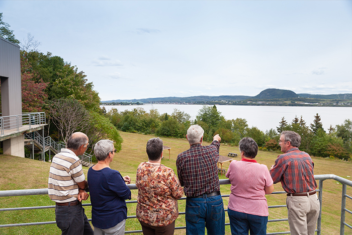 Six adults, three men and three women look in the direction the middle man is pointing with the Restigouche River and New Brunswick's Mount Sugar Loaf in the background.