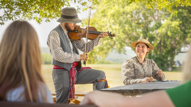 A Parks Canada interpreter in historical costume plays the fiddle while another interpreter sits at a table and watches.