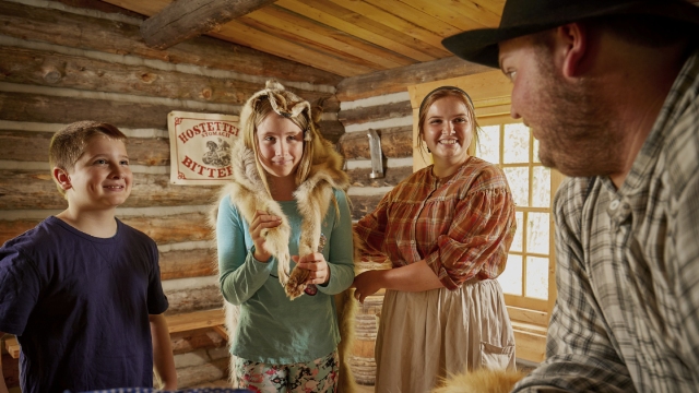 Famille regardant la marchandise offerte dans une cabane de traite métisse, avec l'aide de deux interprètes costumés, au lieu historique national du Fort-Walsh