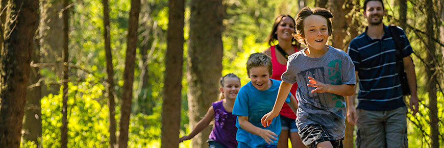 Trois enfants courent sur un sentier dans la forêt devant deux adultes.