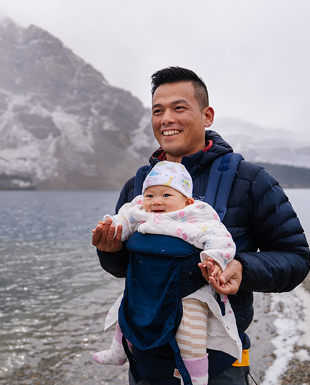 A man with a baby on his chest standing beside a lake