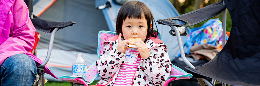 A young girl eats a smore while sitting in a camp chair beside a campfire.