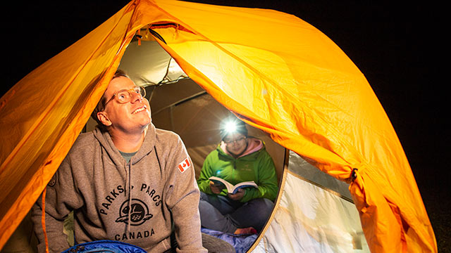 Un jeune homme regarde le ciel par la porte de sa tente et une femme lit un livre au camping de l’arrière-pays du lac Oster, dans le parc national Elk Island