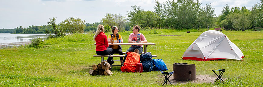 Three backcountry campers sit at a picnic table next to Oster Lake