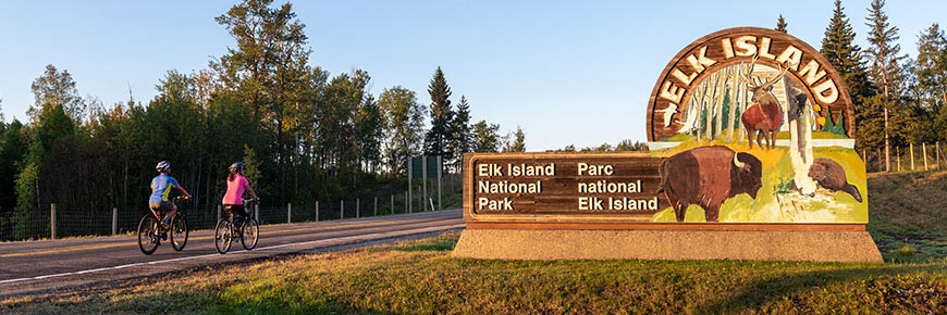 Two cyclists bike past the Elk Island National Park welcome sign.