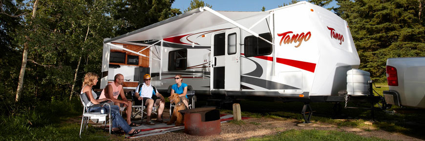A group of four adults and a dog sit around a fire pit in front of an RV.