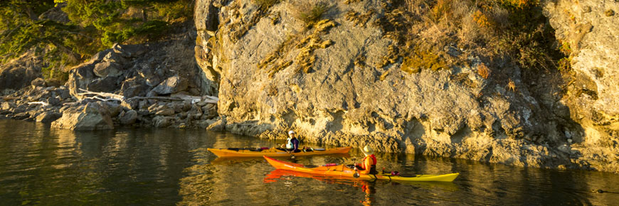 Two kayakers paddle along the shoreline, beneath rocky bluffs in the warm glow of sunset at Pender Island. Gulf Islands National Park Reserve.
