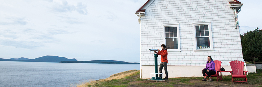 A couple enjoys the viewscope and red chairs at the East Point, Saturna Island Fog Alarm Building.