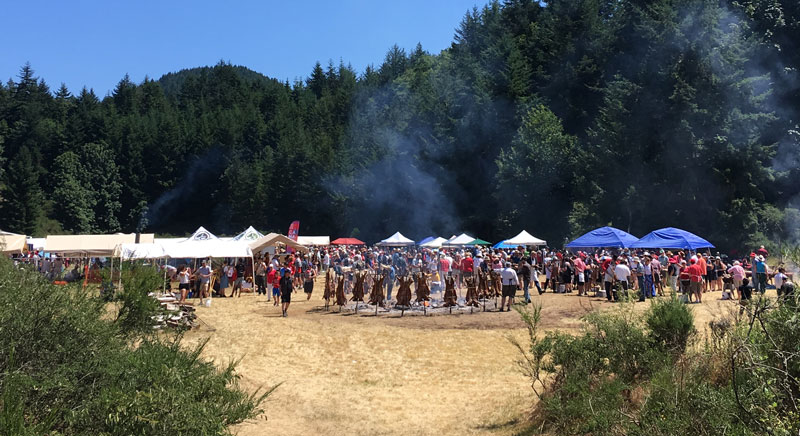 Large canopy tents are up in an open field. People are gathering around a barbecue, and smoke is in the air.