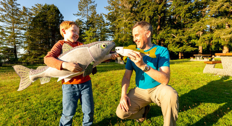 A child holds a large fish replica. His dad stands beside him and holds a smaller fish replica
