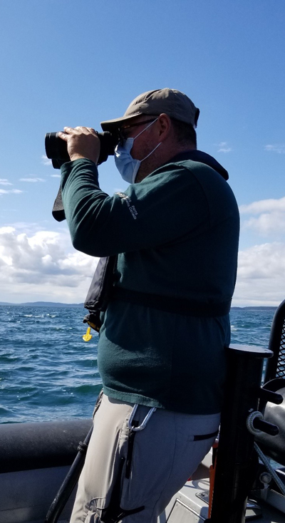 A Parks Canada researcher standing on a vessel in the ocean holds a pair of binoculars to their eyes to look for signs of Southern Resident Killer Whales in the water.