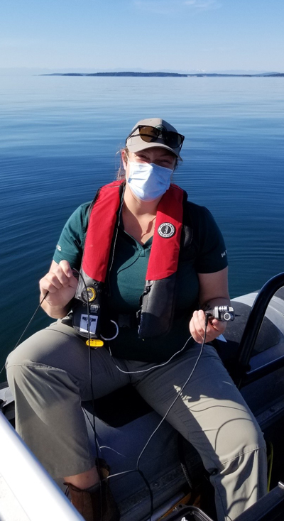 A Parks Canada ecologist wearing a mask smiles at the camera while holding a portable hydrophone unit that has been placed in the ocean.