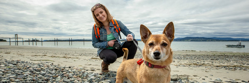 Une femme tenant un chien en laisse sur la plage regarde la caméra.