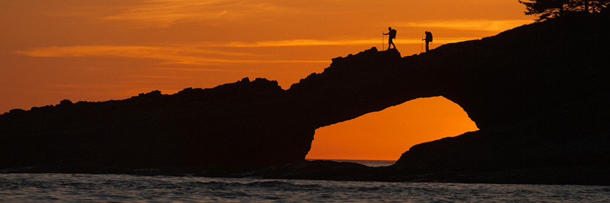 Two hikers in silhouette with packs and poles, cross natural land bridge above water. Orange sunset fills sky in background