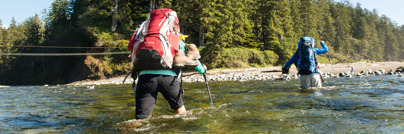 Deux randonneurs portant un sac à dos traversent une rivière à gué, de l’eau aux genoux. On voit des câbles de bac à câble suspendus au-dessus de l’eau à l’arrière-plan.
