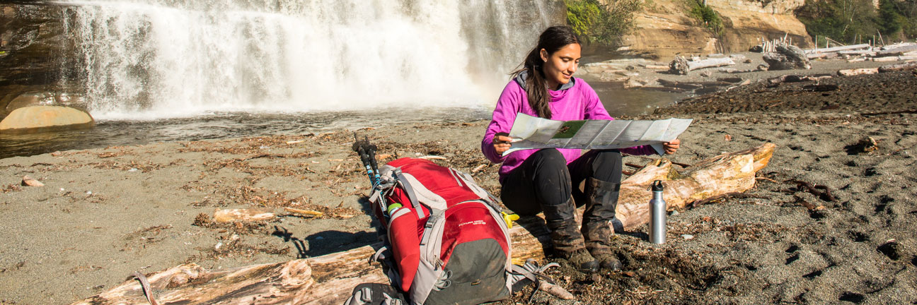 Hiker sits on log consulting map with backpack on ground beside her and waterfall in background