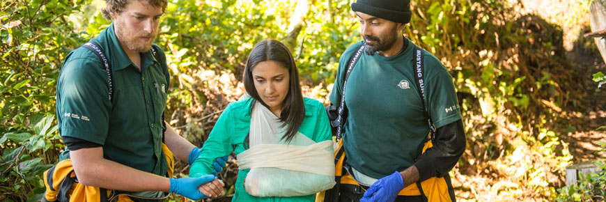 Deux employés de Parcs Canada en uniforme s’occupent d’un randonneur blessé dont le bras est dans une attelle. 