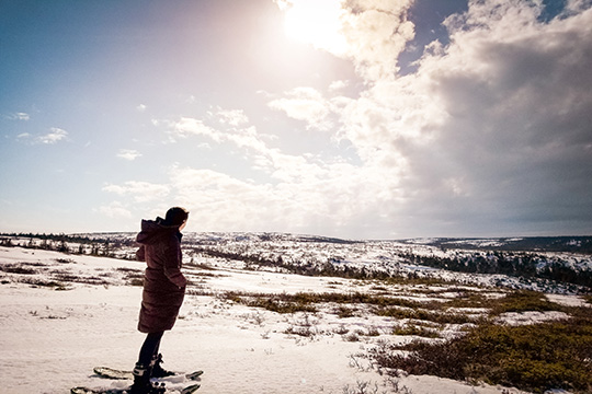 A woman stands on top of a snow covered hill looking out towards the highland's plateau on a partially sunny day