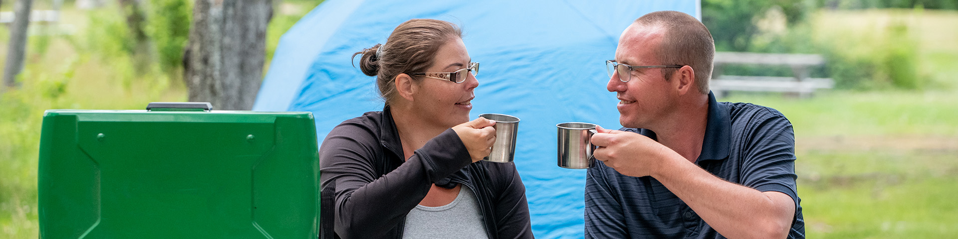 A man and a woman sit next to a camping stove in front of their teal tent, they hold up their steel camping mugs and smile at each other