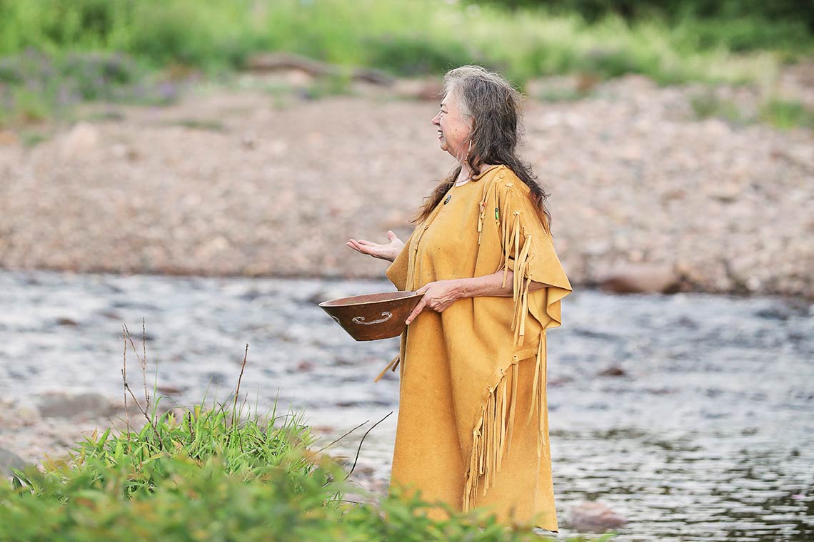 A woman stands by a river holding a wooden bowl and she is wearing traditional Mi'kmaq clothing