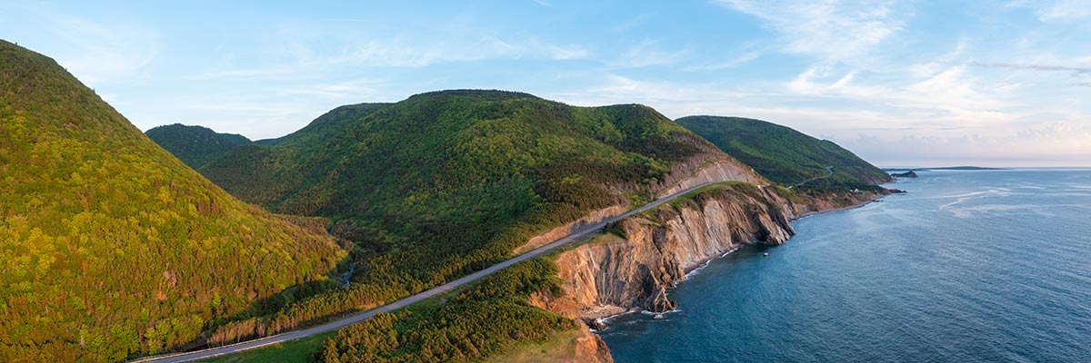 An aerial view of the rolling green mountains and Cabot Trail along rugged cliffs beside the blue ocean