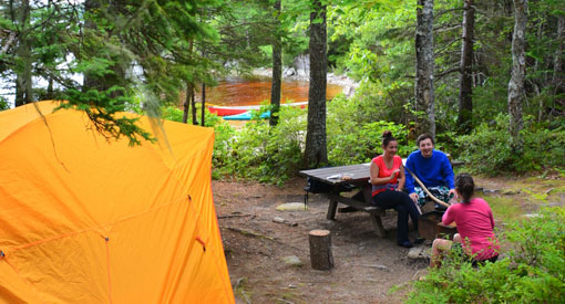 Three campers enjoying a picnic table at a backcountry campsite surrounded by trees with a view of the lake.