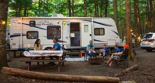 A family sitting at a picnic table and around a campfire with their RV in the background.