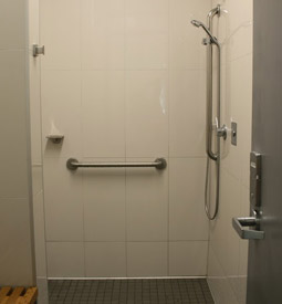 Standard shower with wooden sitting bench, detachable shower wand, dressing hooks, soap rack, and handrail.