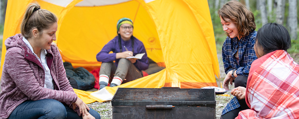 Four people laugh, one from inside a yellow tent, as they cook over a fire pit in a campsite.