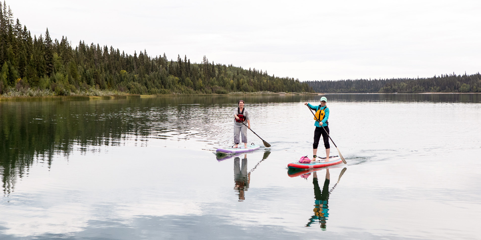 Two women wearing life jackets paddle Pine Lake on stand-up paddleboards. The water is calm and evergreen trees line the shore in the background. 