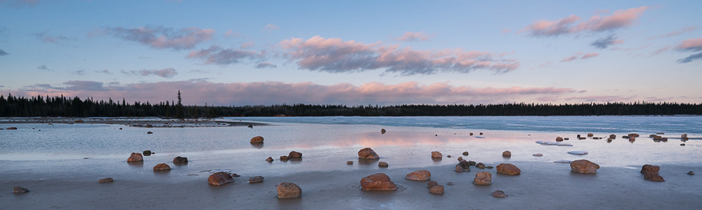 A photo of Grosbeak Lake showing glacial erratics (boulders) strewn across the wet ground. It is dusk and the setting sun turns the sky a hue of pink and blue.