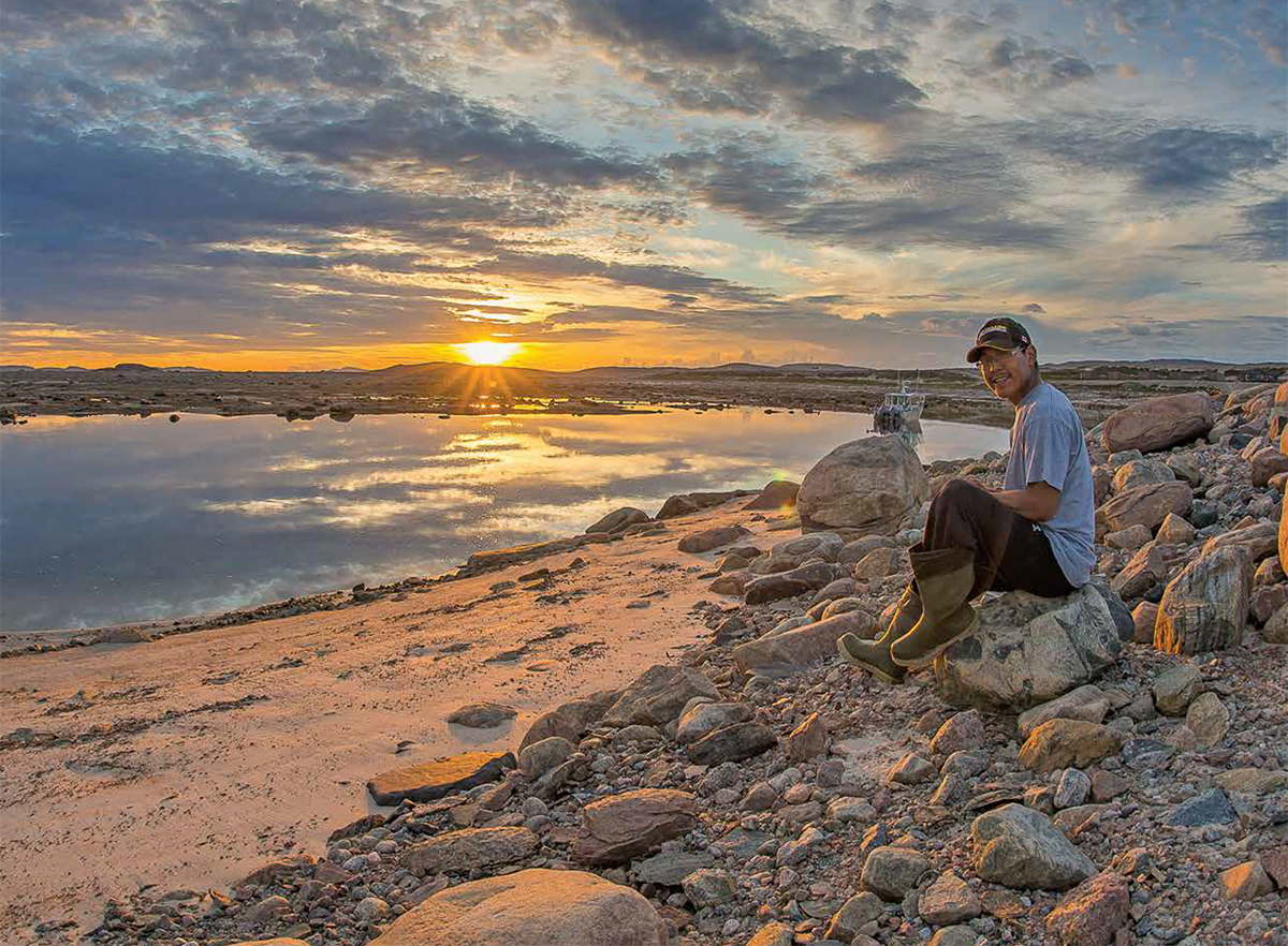 A man sitting on a boulder on a rocky shoreline. A boat is moored behind him. The sun is low on the horizon.