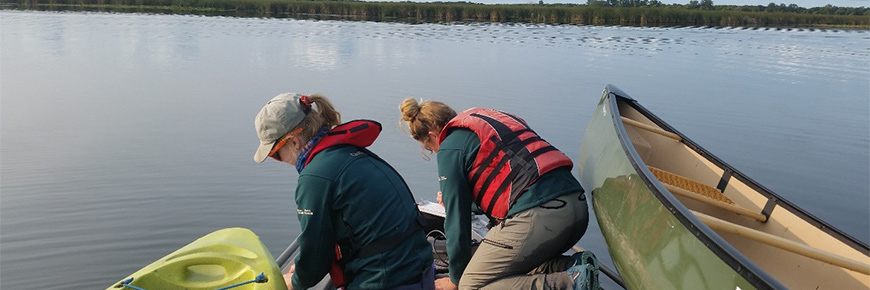 Two members of the resource conservation team conduct water quality monitoring from the bow of a motor boat. The staff member on the left is collecting water samples in bottles, while the member of the right records data. 