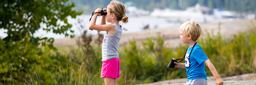 Two children look through binoculars on the shoreline. 