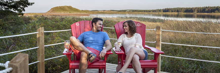 A man and a woman sit in red chairs on the deck of an oTENTik, with Cavendish Beach dunes in the background. 