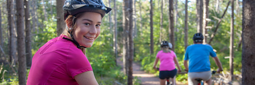 A cyclist looks back and smiles while two other cyclists in the background pedal away, on a wooded trail with green trees.