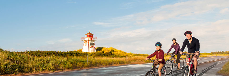 Deux adultes et un enfant, tous portant un casque, passent à vélo devant le phare de Covehead par une journée ensoleillée. 