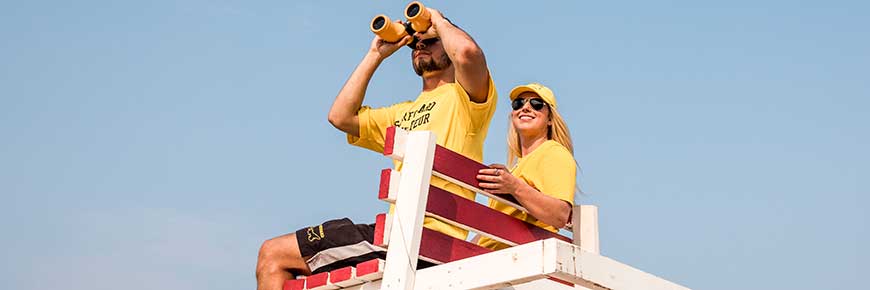 Two surfguards wearing yellow uniforms sitting on a white lifeguard stand. They are both looking off into the distance, one with black binoculars.