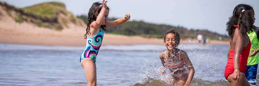 Small children play in shallow water on a sunny day at a PEI National Park beach with dunes in the background. 