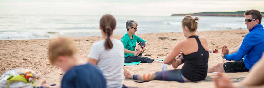 A group yoga class on the beach, with participants sitting and stretching, in front of an instructor with the ocean behind her. 