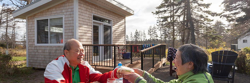 Deux personnes sont assises en souriant à une table de pique-nique devant un Bunkie du camping de Cavendish, avec des chaises Adirondack et un foyer derrière elles.