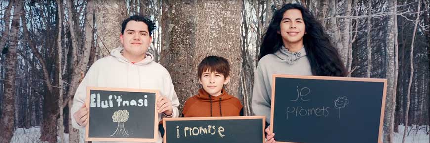 Three male youth hold up signs that say: Elui'tmasi, I Promise, and Je Promets, while standing in a winter forest with lots of snow and trees behind them. . 
