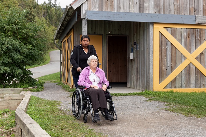 A woman pushes another in a wheelchair. In the background you can see the service building at the Cap-Bon-Ami rest stop.