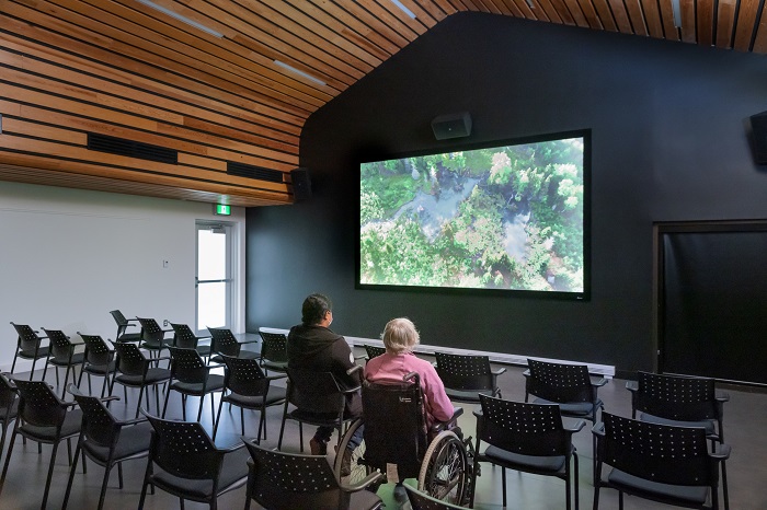 Two women, one in a wheelchair, watch a movie in the screening room at the Visitor Information and Discovery Centre.
