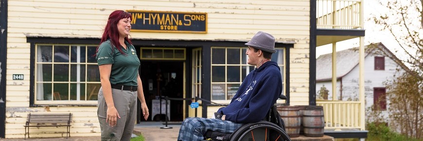 Une employée de Parcs Canada accueille un homme en fauteuil roulant devant le magasin général Hyman and Sons. 