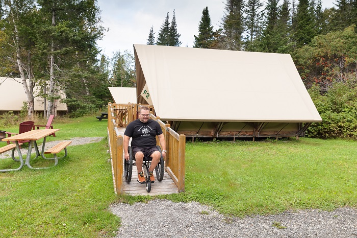 Un homme en fauteuil roulant descend la rampe d'accès qui mène à l'intérieur de la tente oTENTik F2 au camping Petit-Gaspé.