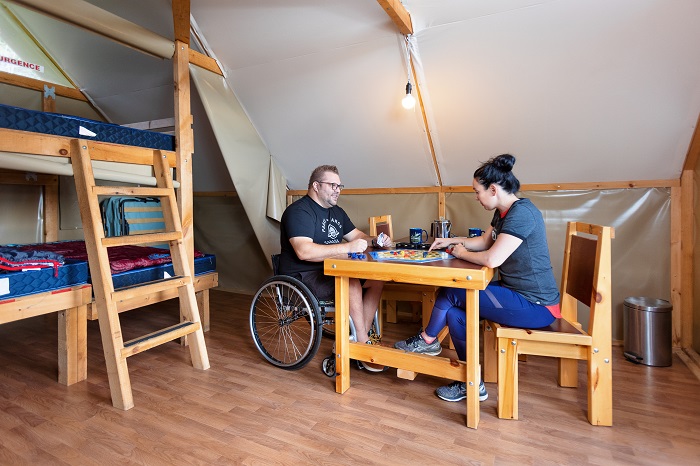 A man in a wheelchair and a woman are seated at a table inside an oTENTik tent. We also see a bed adapted for people with reduced mobility.