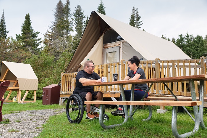 Un homme en fauteuil roulant et une femme sont installés à la table de pique-nique adaptée pour les personnes à mobilité réduite, devant la tente oTENTik F2 du camping Petit-Gaspé. Une rampe permet d'accéder dans la tente. 