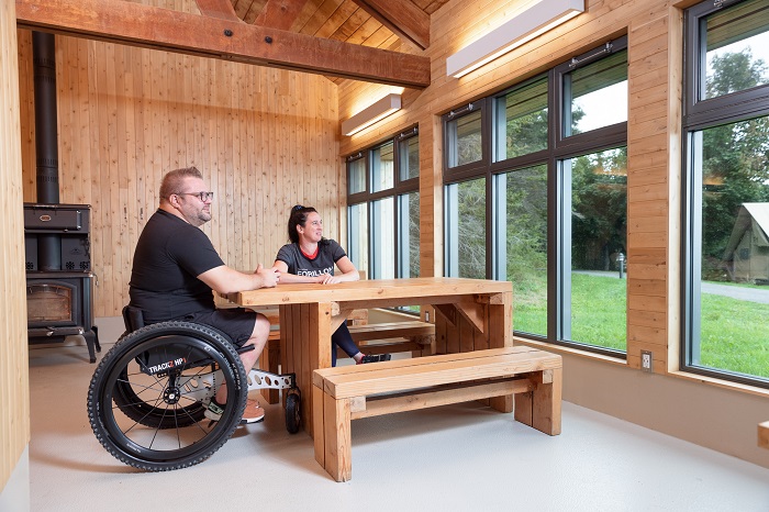 Un homme en fauteuil roulant et une femme sont installés à la table adaptée pour les personnes à mobilité réduite, dans le bâtiment de service de la boucle F du camping Petit-Gaspé.